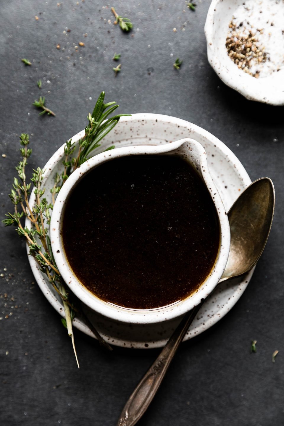 Au jus sauce made with beef drippings fills a small ceramic bowl with a pour spout. The bowl sits atop a matching ceramic plate with fresh herbs and a small spoon resting on top of the plate. A small ceramic pinch bowl filled with kosher salt and black pepper sits alongside the au jus and loose pieces of fresh herbs surround. All items sit atop a dark textured surface.