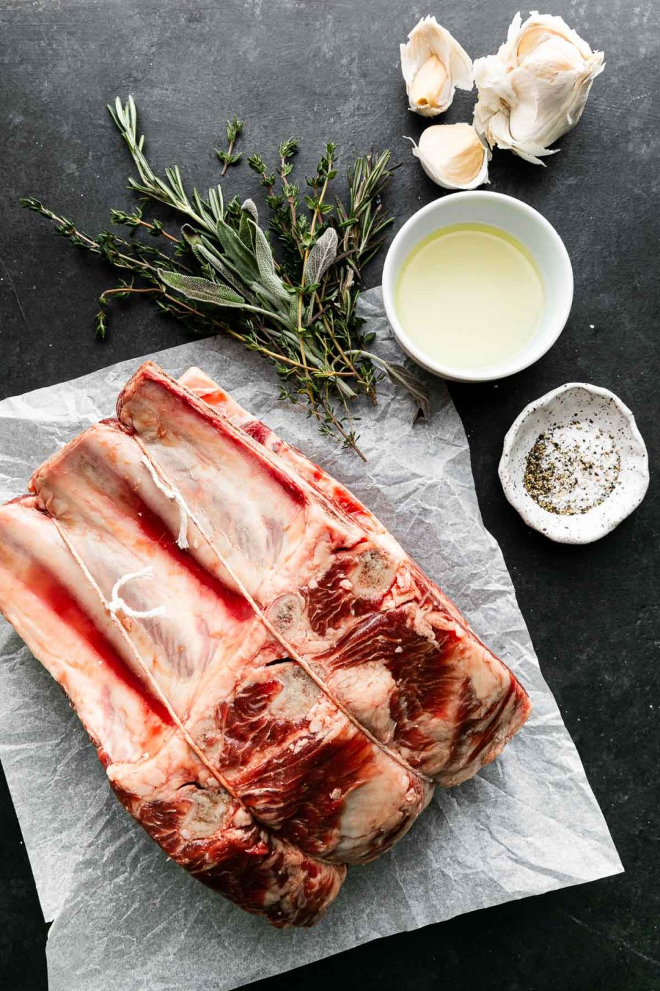 Garlic Herb Crusted Prime Rib ingredients arranged on a dark textured surface: bone-in standing beef rib roast that sits on a sheet of crinkled wax paper, avocado oil, garlic, fresh rosemary, fresh thyme, kosher salt, and ground black pepper.