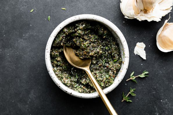Garlic herb rub for Garlic & Herb Prime Rib fills a small ceramic bowl. A Golden spoon rests inside of the bowl and the bowl sits atop a dark textured surface. Cloves of garlic and pieces of fresh thyme rest alongside the bowl.
