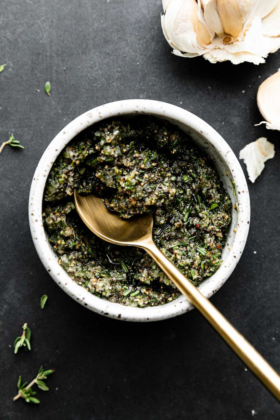 Garlic herb rub for Garlic & Herb Prime Rib fills a small ceramic bowl. A Golden spoon rests inside of the bowl and the bowl sits atop a dark textured surface. Cloves of garlic and pieces of fresh thyme rest alongside the bowl.
