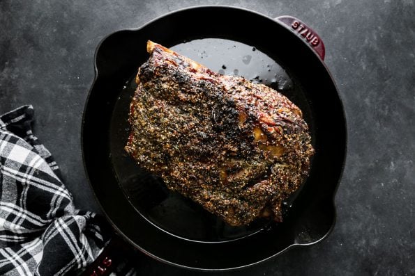 Roasted Prime Rib covered in a Garlic Herb Rub sits inside of a cherry red Staub cast iron Deep Skillet. The skillet sits atop a dark textured surface with a black and white plaid linen napkin resting alongside.