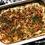 Mom's Loaded Au Gratin Potatoes in a white Staub baking dish sit atop a dark textured surface. A black and white plaid linen napkin rest underneath the baking dish, while a wooden serving spoon and a small ceramic bowl filled with freshly snipped chives rest alongside the baking dish.