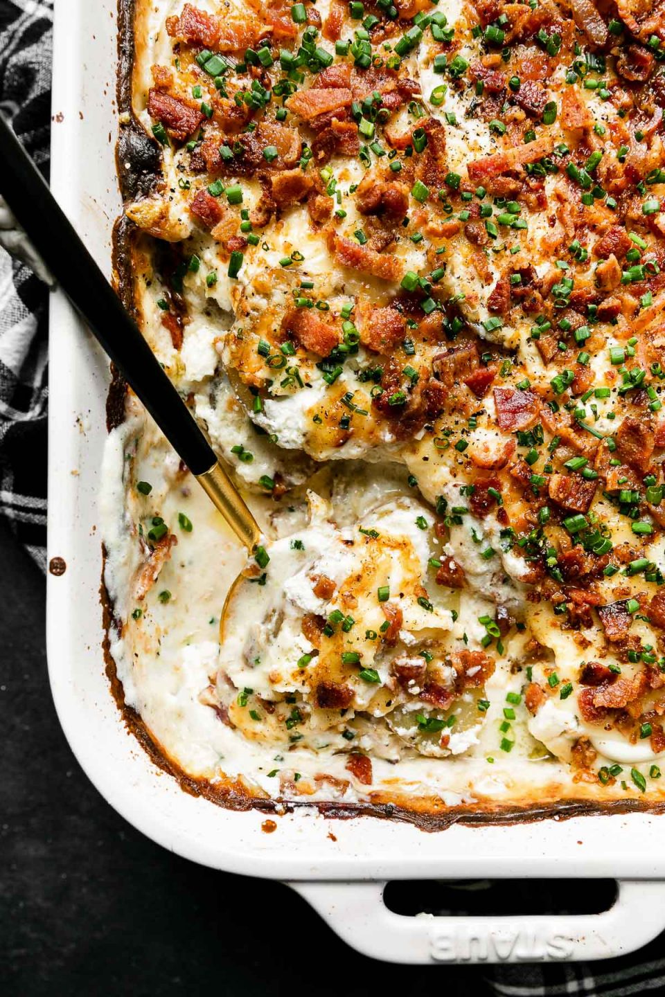 Mom's Loaded Au Gratin Potatoes in a white Staub baking dish sit atop a dark textured surface. A black and white plaid linen napkin rest underneath the baking dish. A black and gold serving spoon rests inside of the baking dish and a portion of the dish has been served.