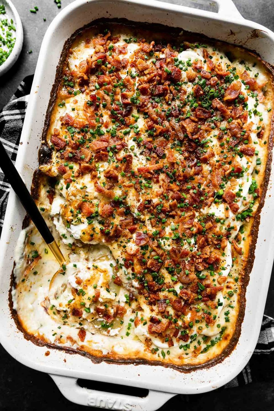 A side angle shot of Mom's Loaded Au Gratin Potatoes in a white Staub baking dish sit atop a dark textured surface. A black and white plaid linen napkin rest underneath the baking dish. A black and gold serving spoon rests inside of the baking dish and a portion of the dish has been served. A small ceramic dish filled with freshly snipped chives rests alongside the baking dish.