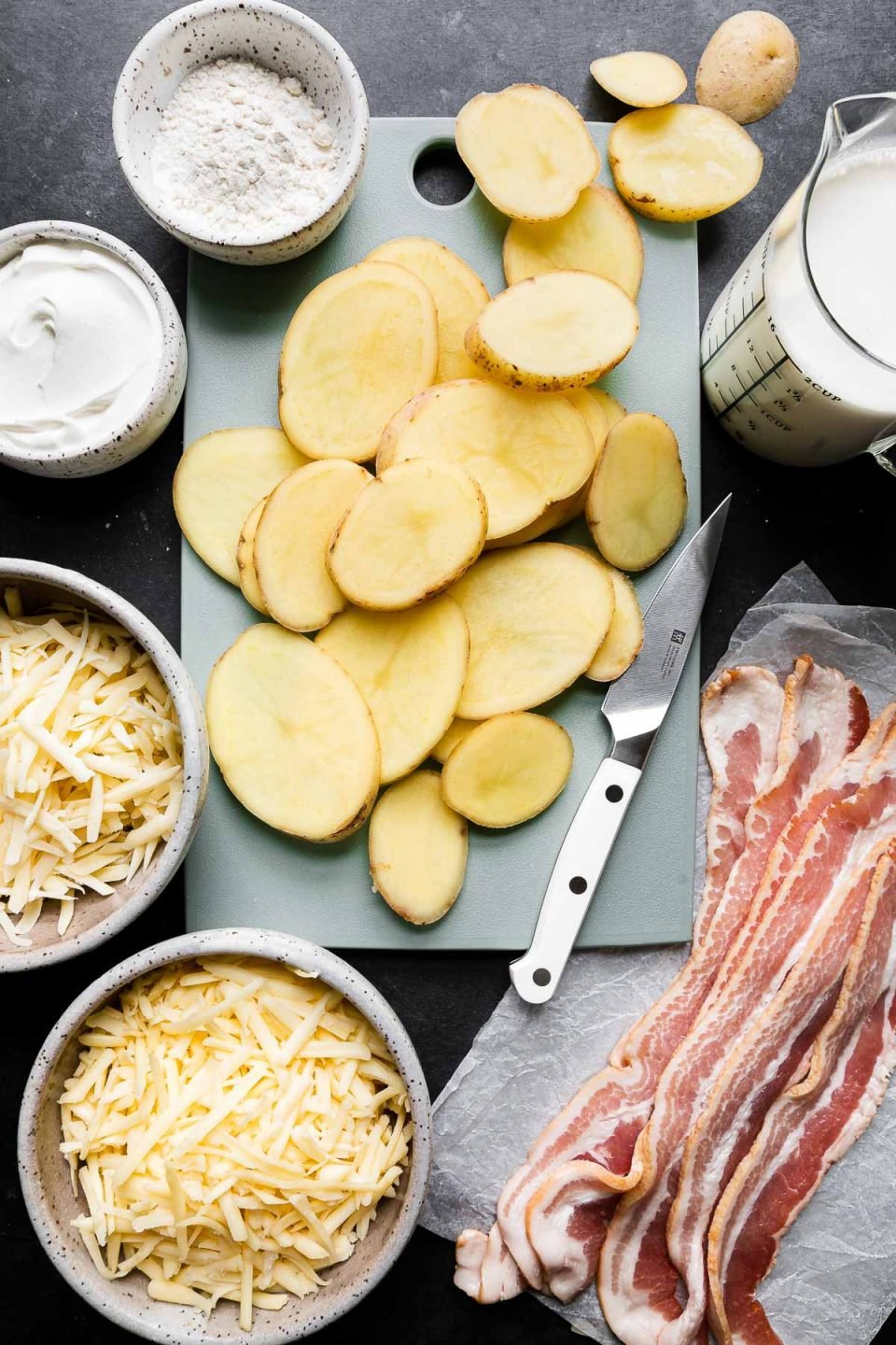 Mom's Loaded Au Gratin Potatoes ingredients arranged on a dark textured surface - sliced Yukon Gold Potatoes sit atop a small blue cutting board, thick-cut bacon, half & half, smoked gouda, gruyère, and sour cream.