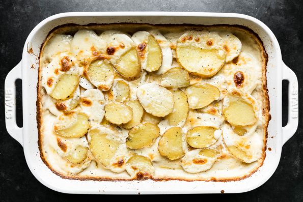 Baked potatoes au gratin in a white Staub baking dish. The baking dish sits atop a dark textured surface.