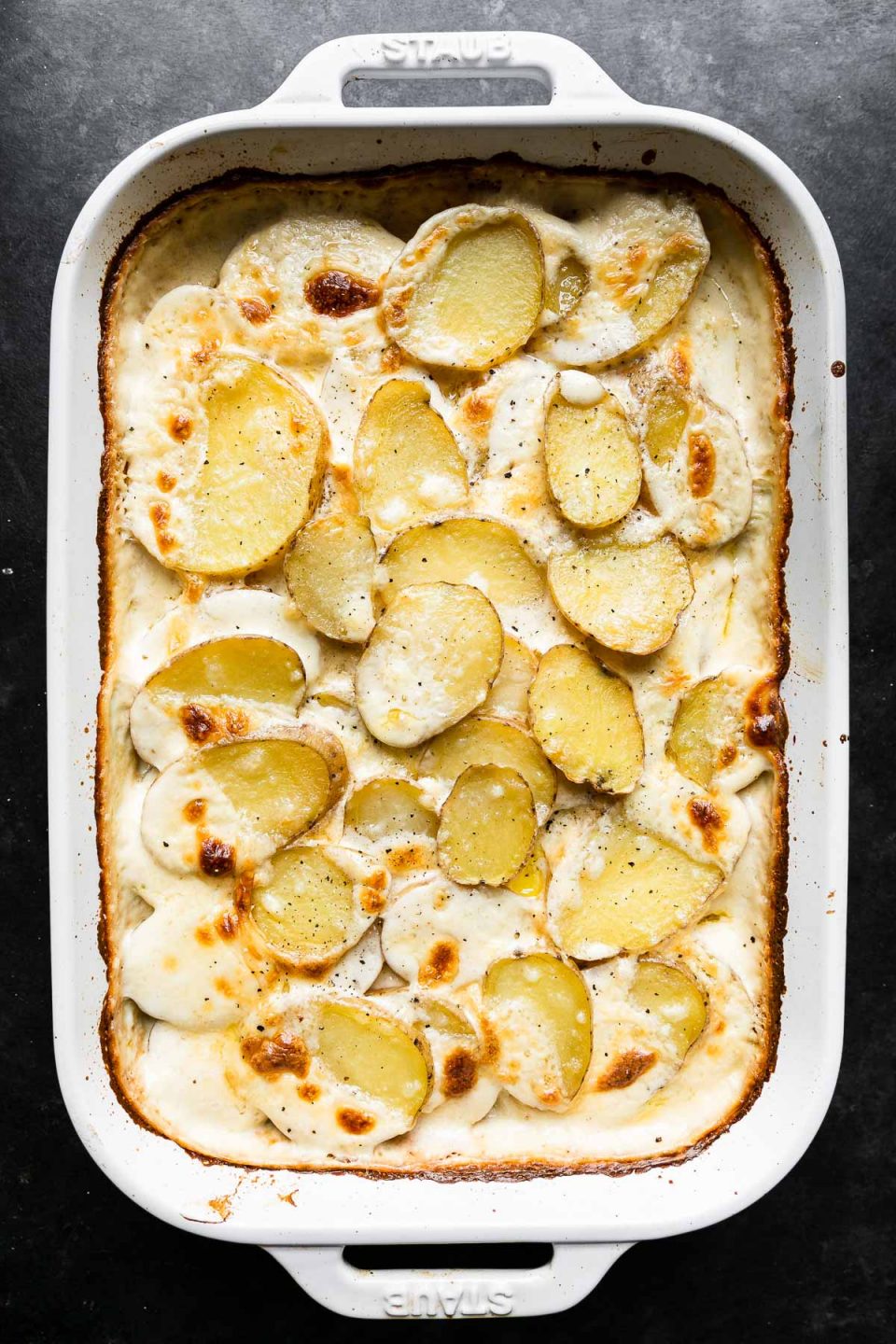 Baked potatoes au gratin in a white Staub baking dish. The baking dish sits atop a dark textured surface.