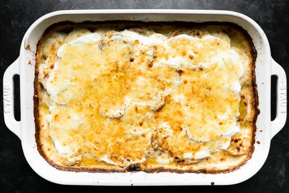 How to make Loaded Au Gratin Potatoes, step 6: Add loaded potato toppings and broil. Baked potatoes au gratin in a white Staub baking dish are layered with sour cream and a sprinkling of gruyère cheese overtop. The gruyère has browned after the dish has been placed underneath the broiler. The baking dish sits atop a dark textured surface.