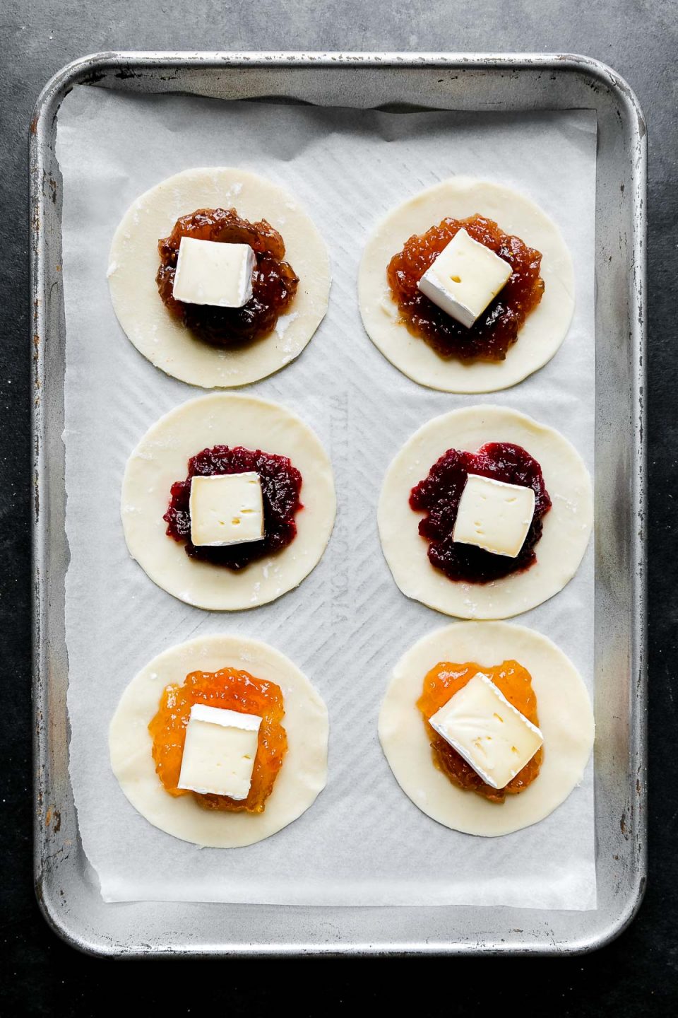 Six 3-inch circles cut out of puff pastry arranged on an aluminum baking sheet lined with parchment paper. Each puff pastry circle has about 1/2 teaspoon of jam or preserve placed in the center with a 1/2-inch piece of brie cheese placed on top to start forming Mini Baked Brie Bites. The baking sheet sits atop a dark textured surface.