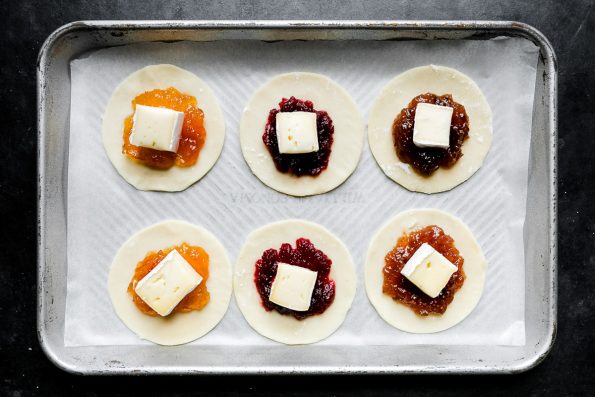 Six 3-inch circles cut out of puff pastry arranged on an aluminum baking sheet lined with parchment paper. Each puff pastry circle has about 1/2 teaspoon of jam or preserve placed in the center with a 1/2-inch piece of brie cheese placed on top to start forming Mini Baked Brie Bites. The baking sheet sits atop a dark textured surface.