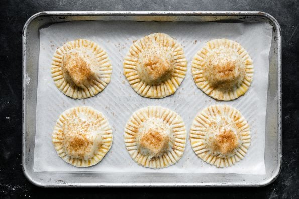 Six assembled Mini Baked Brie Bites arranged on a parchment paper lined baking sheet. The brie bites have been sealed with fork and have crimped edges. The brie bites have been brushed with egg wash and turbinado sugar has been sprinkled over top. The baking sheet sits atop a dark textured surface.