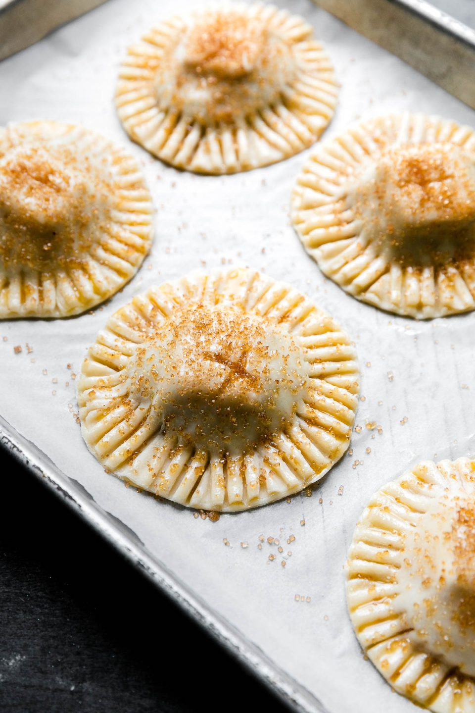 A side angle shot of six assembled Mini Baked Brie Bites arranged on a parchment paper lined baking sheet. The brie bites have been sealed with fork and have crimped edges. The brie bites have been brushed with egg wash and turbinado sugar has been sprinkled over top. The baking sheet sits atop a dark textured surface.