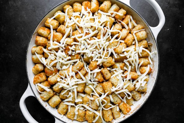 An assembled Truffled Steak Tater Tot Hotdish fills a heavy-bottomed pan. A layer of baked & crispy tater tots rest on top witha. sprinkle of truffle cheese over top the tater tots as well prior to baking. The pan sits atop a black textured surface.