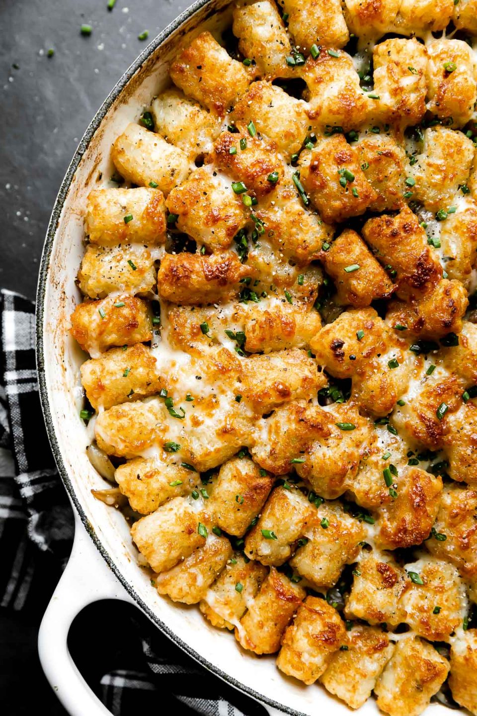 A finished Truffled Steak Tater Tot Hotdish made in a white heavy-bottomed pan sits atop a dark textured surface. A black and white plaid linen napkin rests alongside the pan and the hotdish has been garnished with freshly chopped herbs.