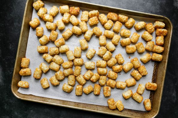Golden brown baked tater tots arranged in a single layer rest atop a parchment lined baking sheet. The baking sheet sits atop a dark textured surface.