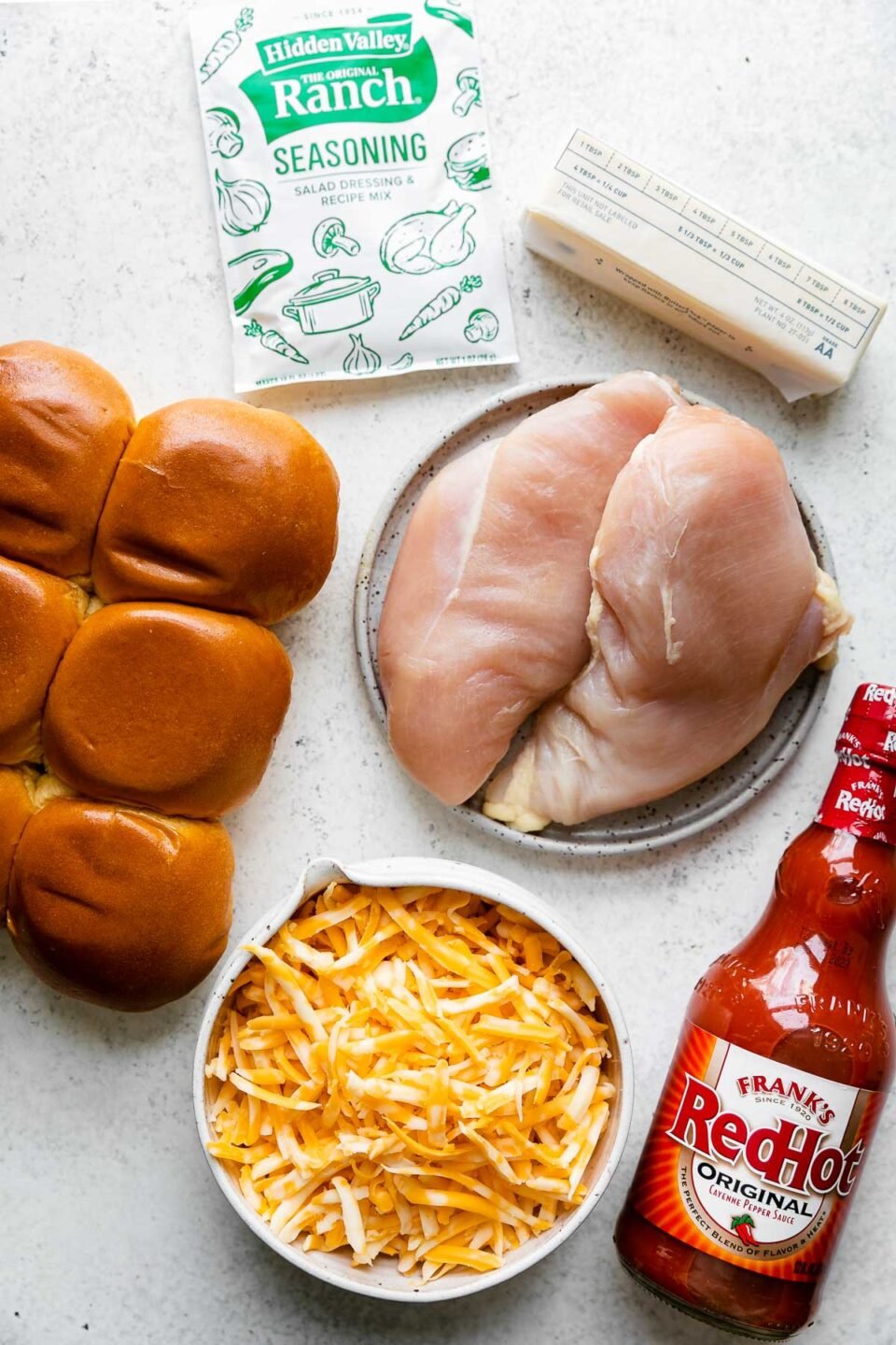 Buffalo Chicken Slider ingredients arranged on a white textured surface: unsalted butter, boneless, skinless chicken breasts, ranch seasoning, Frank's RedHot Original Cayenne Pepper Sauce, dinner rolls, Colby jack cheese.