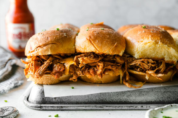 Buffalo Chicken Sliders on brioche buns. The sliders are placed atop a piece of parchment paper atop an upside down aluminum baking sheet. The baking sheet sits atop a creamy white textured surface. A bottle of Frank's RedHot Sauce, a beer cap bottle, a small ceramic bowl filled with ranch dressing, and loose freshly snipped chives surround the sliders.