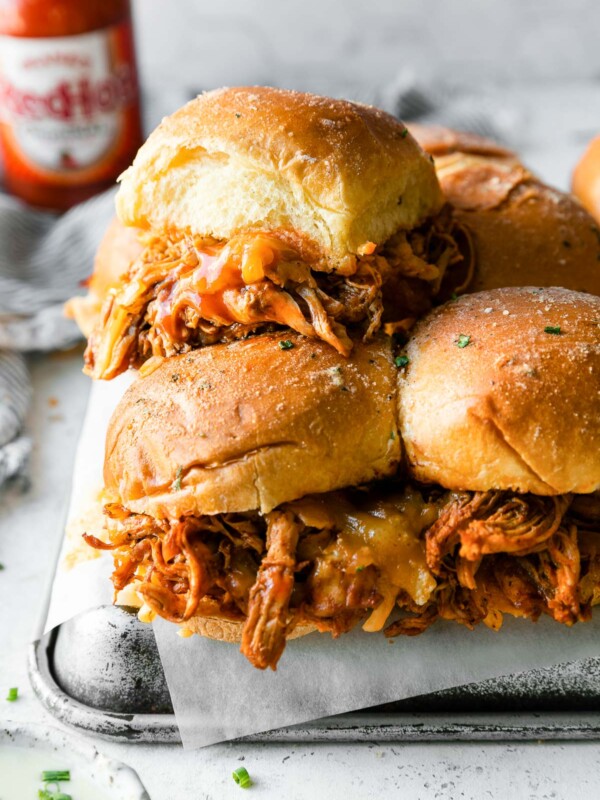 Buffalo Chicken Sliders on brioche buns are stacked atop a piece of parchment paper and resting atop an upside down aluminum baking sheet. The baking sheet sits atop a creamy white textured surface. A blue and white striped linen napkin, a bottle of Frank's RedHot sauce, a small ceramic bowl filled with ranch dressing, and loose freshly snipped chives surround the sliders.