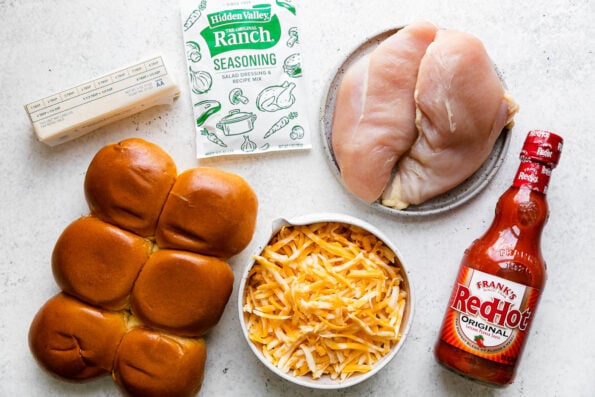 Buffalo Chicken Slider ingredients arranged on a white textured surface: unsalted butter, boneless, skinless chicken breasts, ranch seasoning, Frank's RedHot Original Cayenne Pepper Sauce, dinner rolls, Colby jack cheese.