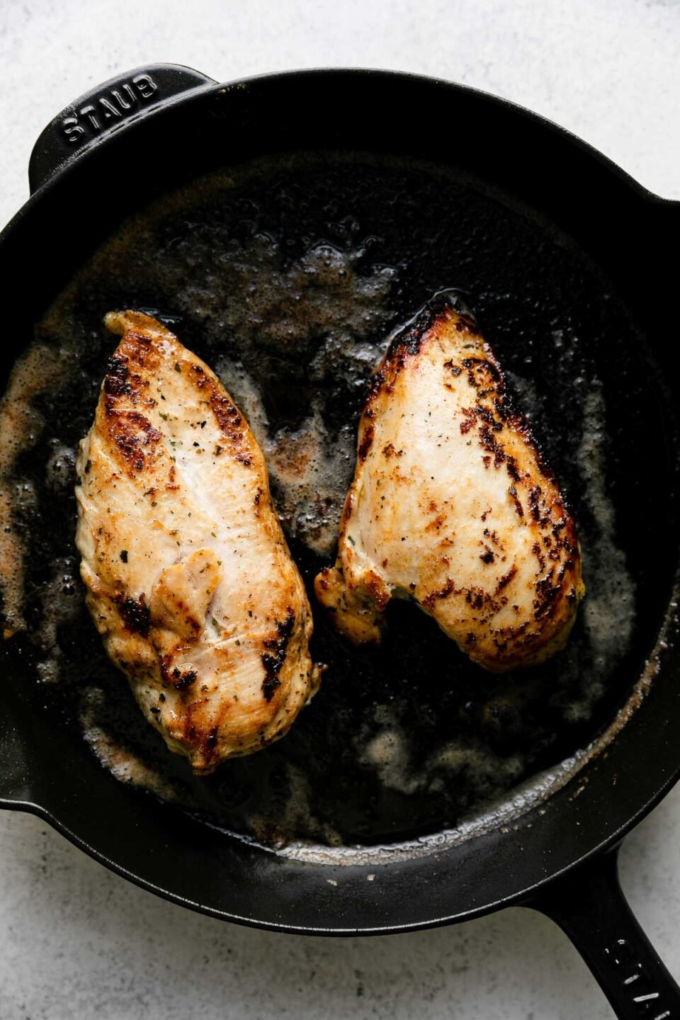 Two boneless, skinless chicken breasts are seared in butter in a black Staub cast iron skillet. The skillet sits atop a creamy white textured surface.