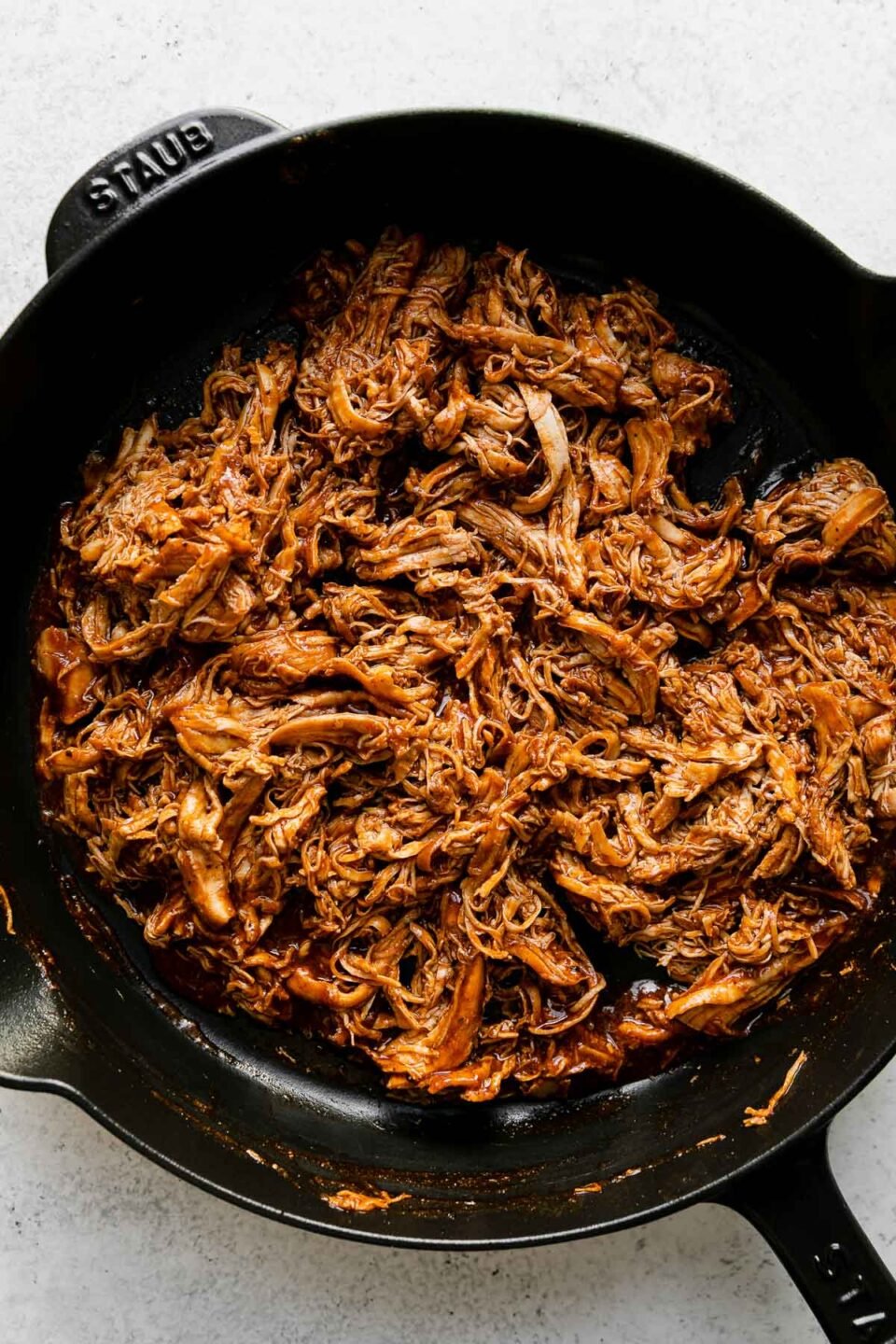 Shredded chicken mixed with buffalo sauce fills a black Staub cast iron skillet. The skillet sits atop a creamy white textured surface.