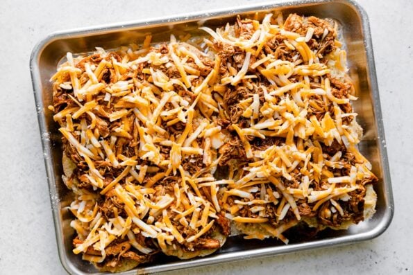 How to make Buffalo Chicken Sliders, step 5: Buffalo Chicken Slider Assembly. The bottom portion of a slab of dinner rolls is topped with Colby jack cheese, shredded buffalo chicken, then another layer of Colby jack cheese. The sliders are assembled atop a baking sheet and the baking sheet sits atop a creamy white textured surface.