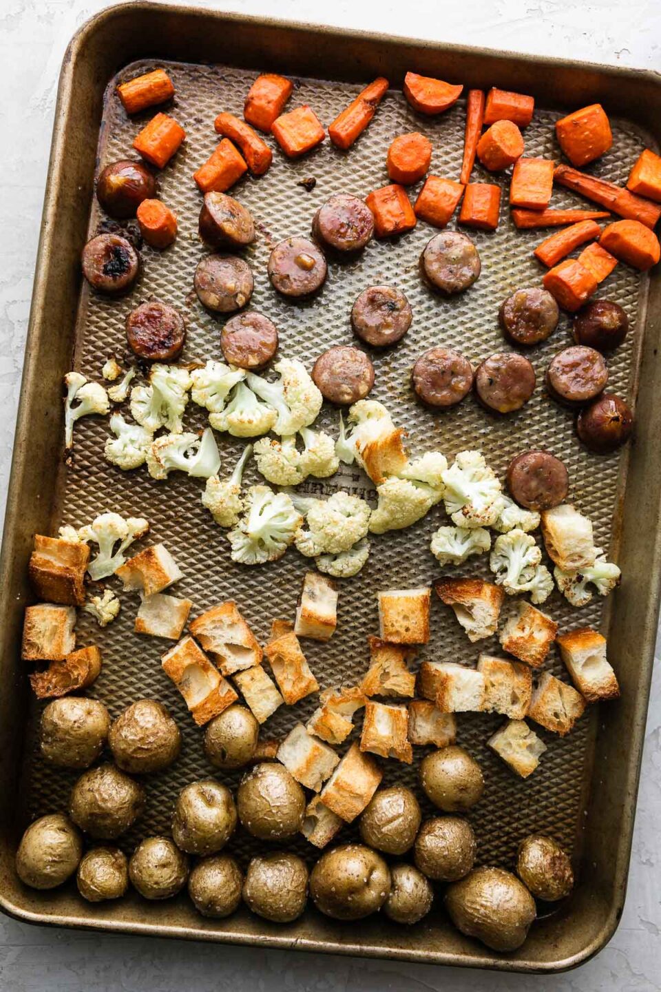 How to make Fondue for Two, Steps 2 & 3, roast the fondue dippers. Roasted and golden brown carrots, baby potatoes, cauliflower, bread, and chicken sausage, cut into bite-sized pieces, are all arranged on a baking sheet. The baking sheet sits atop a creamy white textured surface.