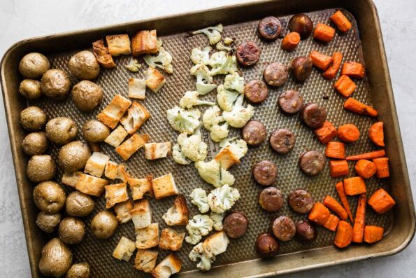 How to make Fondue for Two, Steps 2 & 3, roast the fondue dippers. Roasted and golden brown carrots, baby potatoes, cauliflower, bread, and chicken sausage, cut into bite-sized pieces, are all arranged on a baking sheet. The baking sheet sits atop a creamy white textured surface.