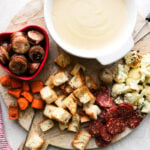 A cheese fondue for two spread is set up on a wooden cheese board. Cheese fondue fills a white fondue pot or caquelon which rests atop the cheese board with bite-sized pieces of roasted carrots, bread, cauliflower, potatoes, chicken sausage, and mini salami for dipping surrounding. Two fondue forks rest atop the cheese board which sits atop a creamy white textured surface. A bottle of Exquisite Collection Sauvignon Blanc, a red and white striped linen napkin, and two glasses of white wine surround the cheese board.