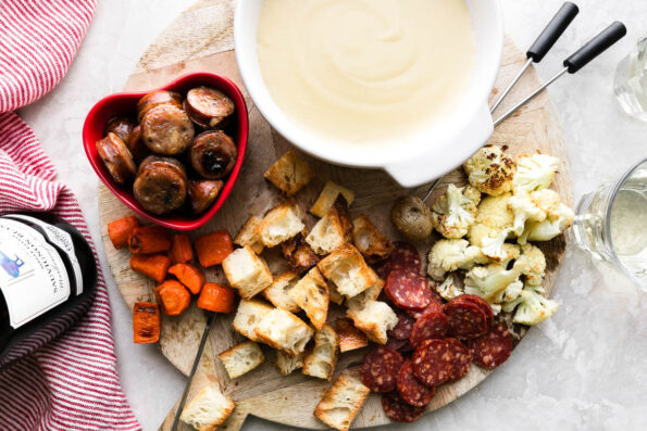 A cheese fondue for two spread is set up on a wooden cheese board. Cheese fondue fills a white fondue pot or caquelon which rests atop the cheese board with bite-sized pieces of roasted carrots, bread, cauliflower, potatoes, chicken sausage, and mini salami for dipping surrounding. Two fondue forks rest atop the cheese board which sits atop a creamy white textured surface. A bottle of Exquisite Collection Sauvignon Blanc, a red and white striped linen napkin, and two glasses of white wine surround the cheese board.