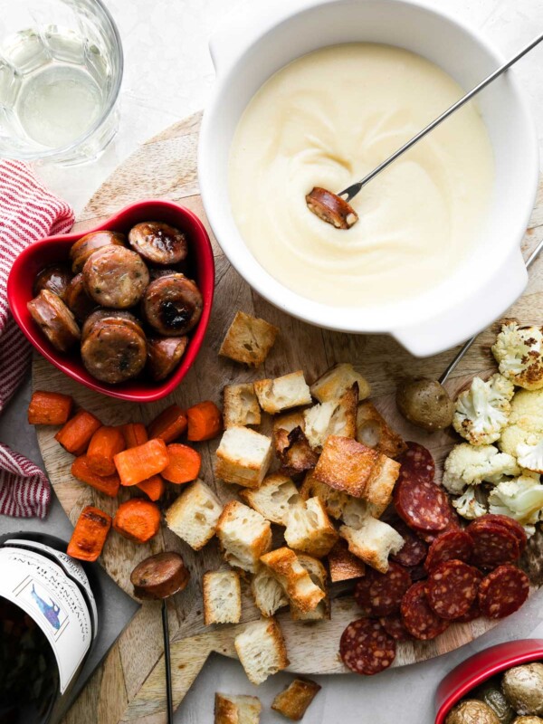 An overhead shot of a cheese fondue for two spread is set up on a wooden cheese board. Cheese fondue fills a white fondue pot or caquelon which rests atop the cheese board with bite-sized pieces of roasted carrots, bread, cauliflower, potatoes, chicken sausage, and mini salami for dipping surrounding. One fondue fork holding a piece of chicken sausage is dipped into the fondue while another rests atop the cheese board which sits atop a creamy white textured surface. A bottle of Exquisite Collection Sauvignon Blanc, a red and white striped linen napkin, a glass of white wine, and a red heart shaped bowl filled with roasted baby potatoes rest alongside the cheese board.