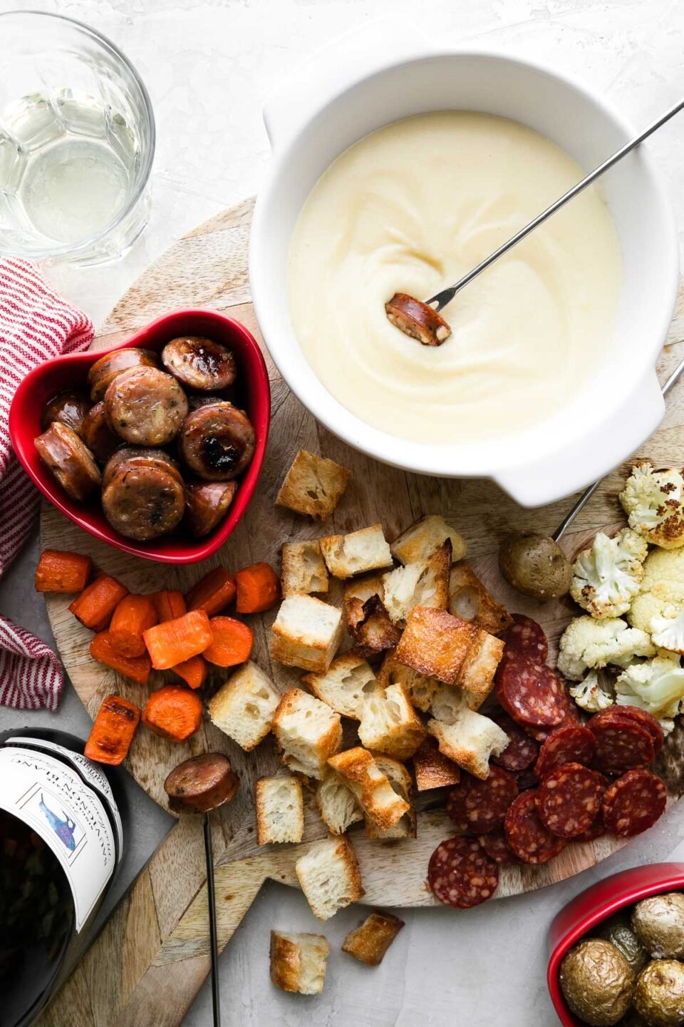 An overhead shot of a cheese fondue for two spread is set up on a wooden cheese board. Cheese fondue fills a white fondue pot or caquelon which rests atop the cheese board with bite-sized pieces of roasted carrots, bread, cauliflower, potatoes, chicken sausage, and mini salami for dipping surrounding. One fondue fork holding a piece of chicken sausage is dipped into the fondue while another rests atop the cheese board which sits atop a creamy white textured surface. A bottle of Exquisite Collection Sauvignon Blanc, a red and white striped linen napkin, a glass of white wine, and a red heart shaped bowl filled with roasted baby potatoes rest alongside the cheese board.