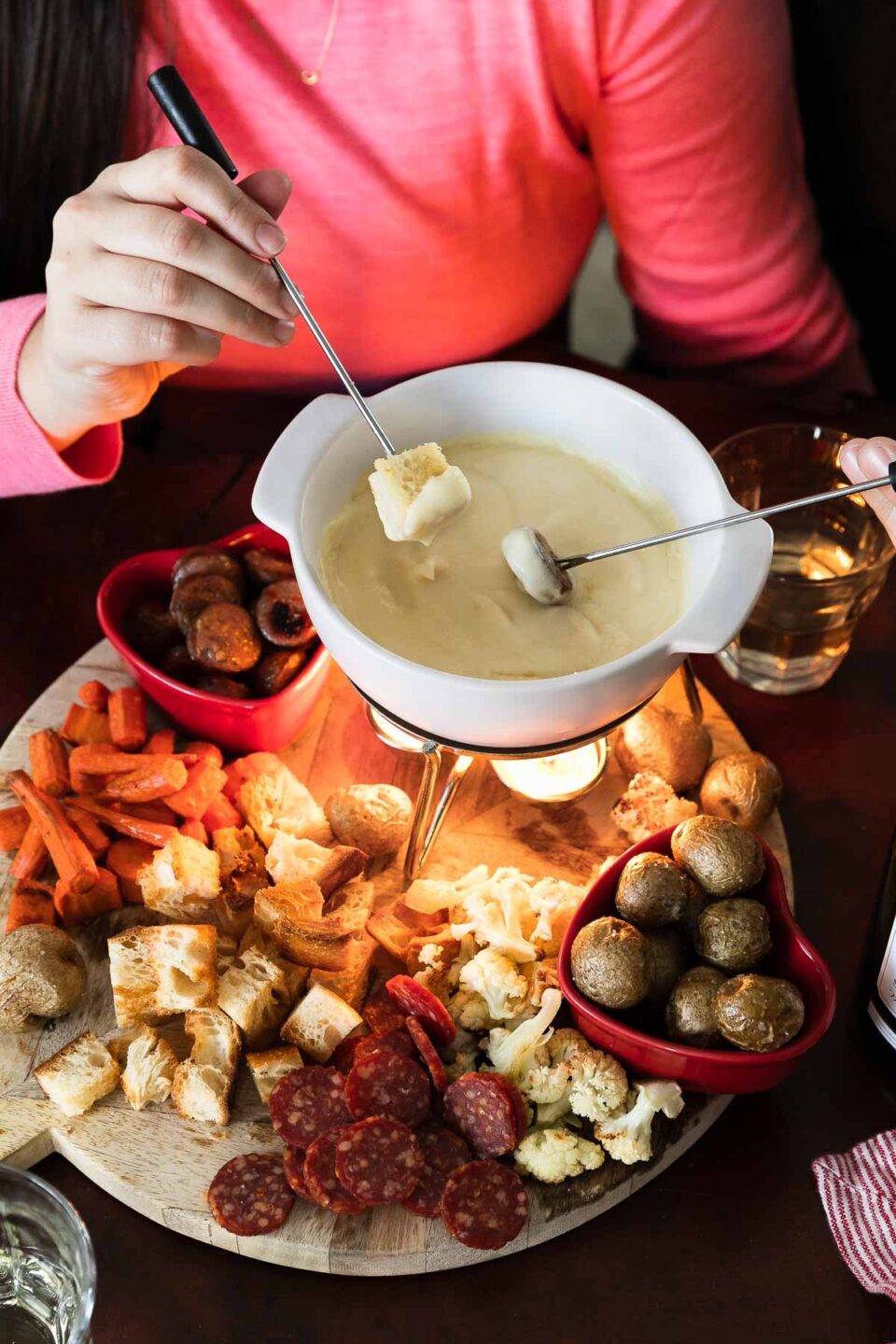 An overhead shot of a cheese fondue for two spread is set up on a wooden cheese board. Cheese fondue fills a white fondue pot or caquelon which rests atop the cheese board with bite-sized pieces of roasted carrots, bread, cauliflower, potatoes, chicken sausage, and mini salami for dipping surrounding. The cheese board sits atop a wood surface. A woman's hand holds a fondue fork with a piece of toasted bread attached and dips it into the fondue pot. While a man holds another fondue fork with a piece of chicken sausage on the end also dipping into the pot. Two glasses of white wine, and a red and white striped linen napkin rest alongside the cheese board.