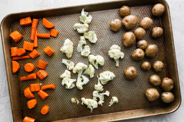 Carrots, baby potatoes, and cauliflower cut into bite-sized pieces arranged on a baking sheet for roasting. The baking sheet sits atop a creamy white textured surface.