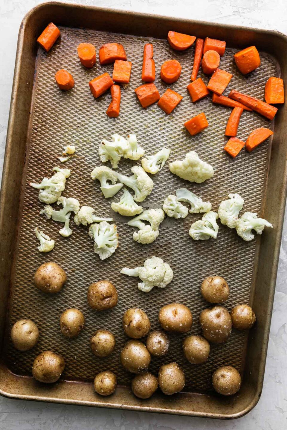 Carrots, baby potatoes, and cauliflower cut into bite-sized pieces arranged on a baking sheet for roasting. The baking sheet sits atop a creamy white textured surface.