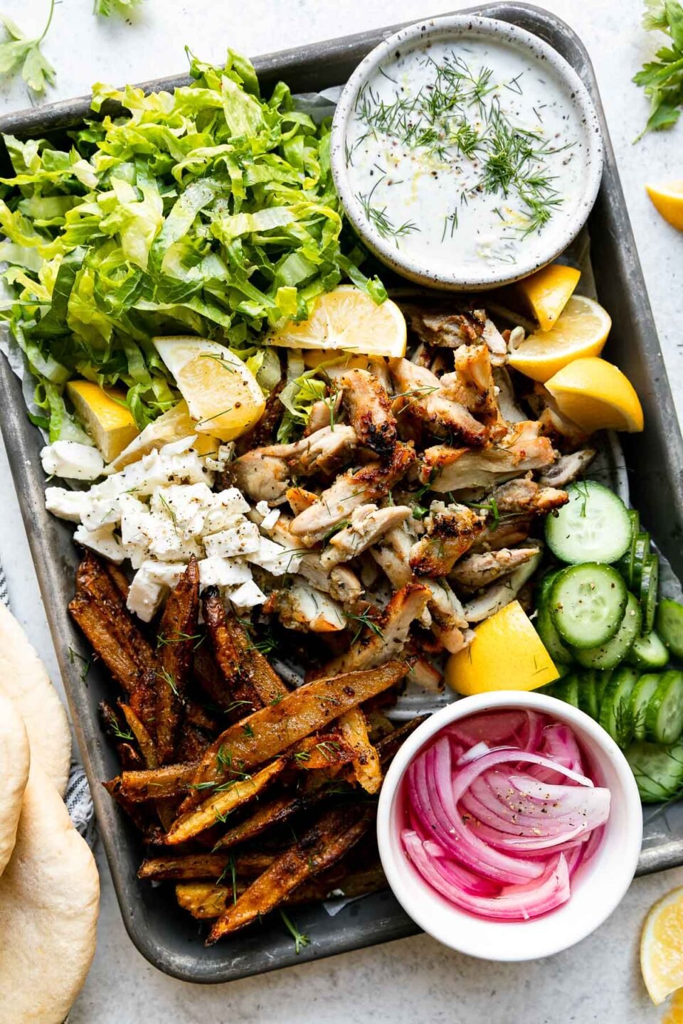 A small baking sheet is filled with all of the fixings to make Lemony Greek Chicken Pitas: shredded lettuce, sliced cucumber, oven-roasted Greek fries, sliced lemony Greek chicken, crumbled feta, lemon wedges, a bowl filled with tzatziki-ish herbed yogurt sauce, and a small pinch bowl filled with pickled red onions. Warm pitas rest alongside the sheet pan wrapped in a blue and white striped linen napkin along with lemon wedges and loose fresh herbs that surround the baking sheet. All items rest atop a creamy white textured surface.