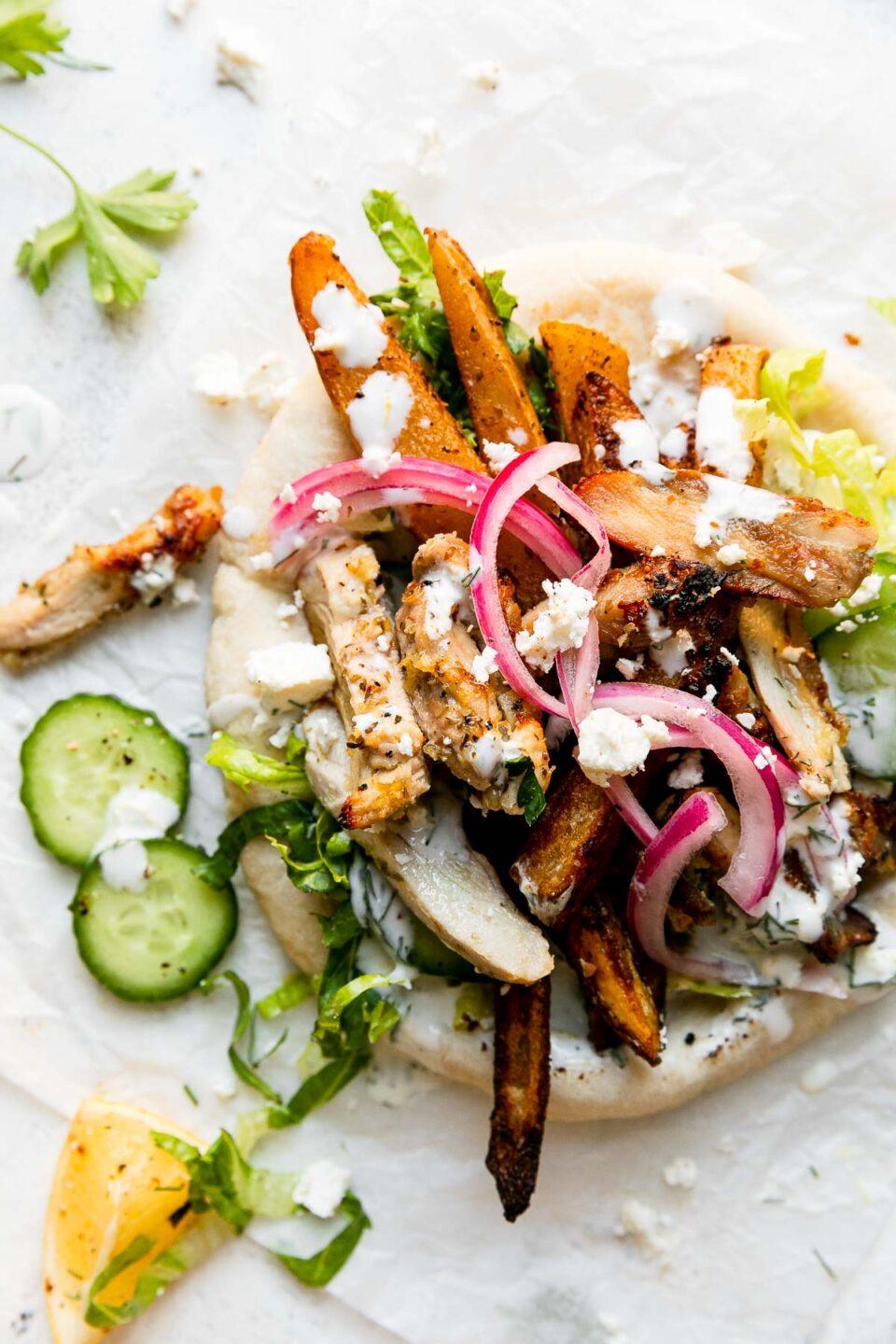 An assembled Greek Chicken Pita rests atop a crumpled sheet of white parchment paper. The pita has been piled high with sliced lemony Greek chicken, oven-roasted Greek fries, shredded lettuce, crumbled feta, sliced cucumber, pickled red onion, and a drizzle of tzatziki-ish herbed yogurt sauce. Surrounding the pita is loose fresh herbs, a few slices of cucumber, a lemon wedge, and crumbled feta. All items sit atop a creamy white textured surface.