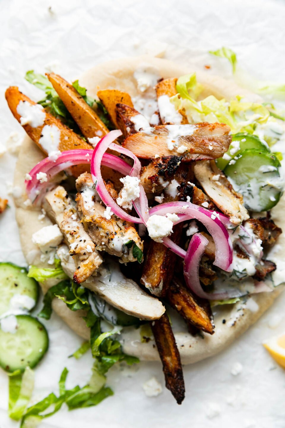 An overhead shot of an assembled Greek Chicken Pita that rests atop a crumpled sheet of white parchment paper. The pita has been piled high with sliced lemony Greek chicken, oven-roasted Greek fries, shredded lettuce, crumbled feta, sliced cucumber, pickled red onion, and a drizzle of tzatziki-ish herbed yogurt sauce. Surrounding the pita is a few slices of cucumber, a lemon wedge, and crumbled feta. All items sit atop a creamy white textured surface.