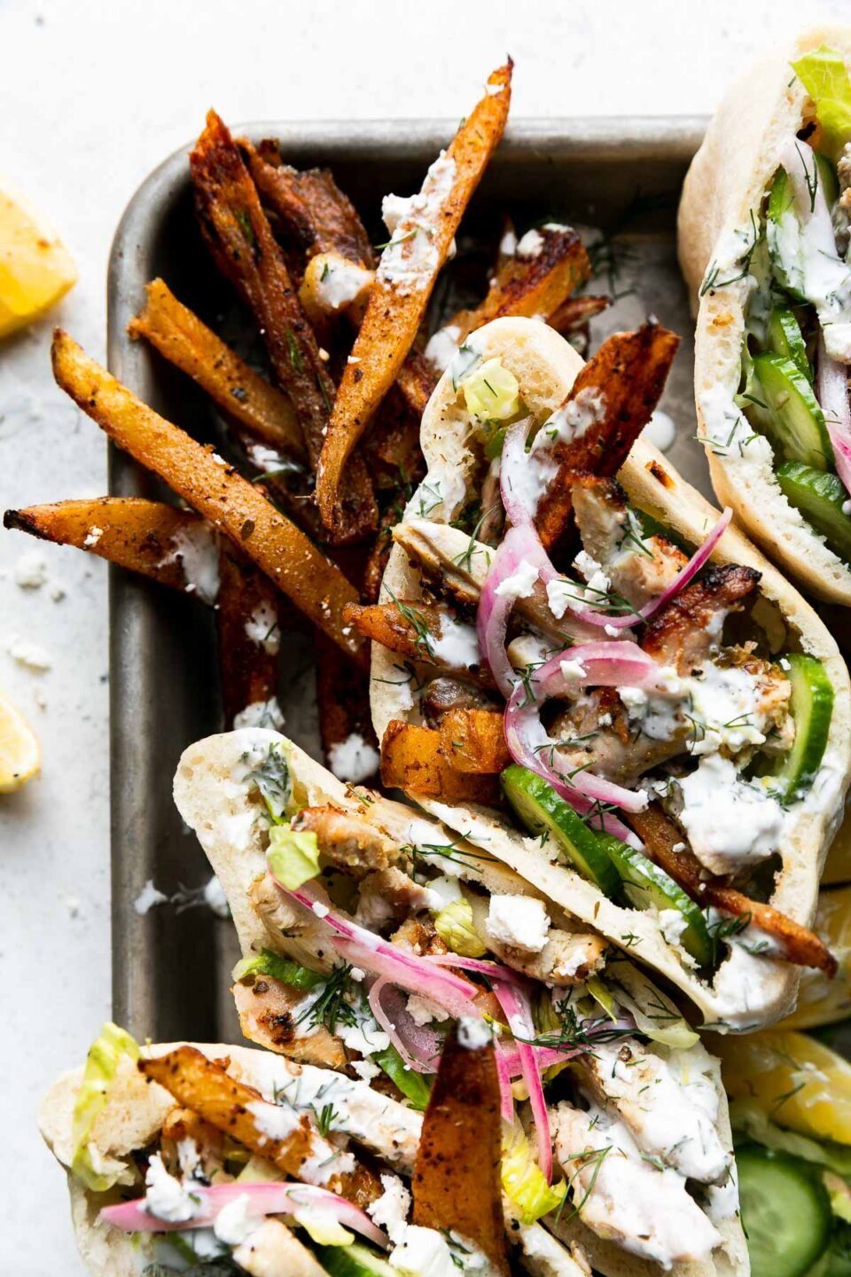 Four Lemony Greek Chicken Pita sandwiches arranged on a small baking sheet alongside a small pile of oven-roasted Greek fries, lemon wedges, and sliced cucumber. Lemon wedges and crumbled feta surround the baking sheet and all items sit atop a creamy white textured surface.