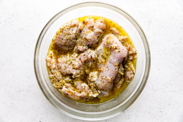 Boneless skinless chicken thighs rest in a glass mixing bowl of Lemony Greek marinade. The bowl sits atop a creamy white textured surface.