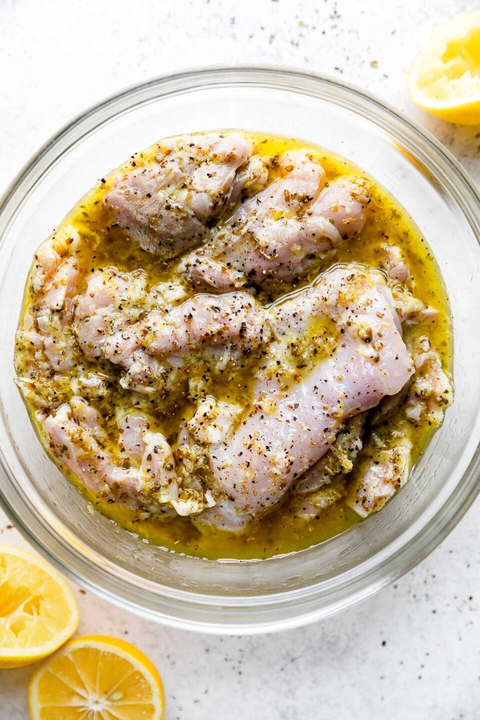 Boneless skinless chicken thighs rest in a glass mixing bowl of Lemony Greek marinade. The bowl sits atop a creamy white textured surface. Halved lemons rest on the surface surrounding the bowl with the exposed fruit facing upwards.