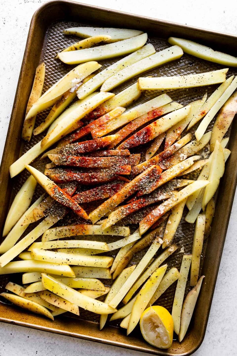 Gold potatoes sliced into 1/3-inch fries are arranged on a small baking sheet. The potatoes have been prepared for roasting with a drizzle of olive oil, and seasoned with lemon juice, dried oregano, smoked paprika, & garlic powder over top. One lemon halved rests along with the sliced potato on the baking sheet and the baking sheet sits atop a creamy white textured surface.