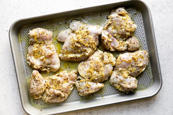 Six marinated boneless skinless chicken thighs arranged on a small baking sheet. The baking sheet sits atop a creamy white textured surface.