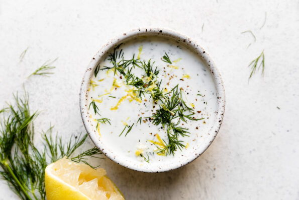 A small white speckled ceramic bowl is filled with tzatziki-ish herbed yogurt drizzle. The yogurt drizzle is garnished with fresh dill, lemon zest, and ground black pepper. The bowl sits atop a creamy white textured surface with more fresh dill and a single halved lemon surrounding the bowl.