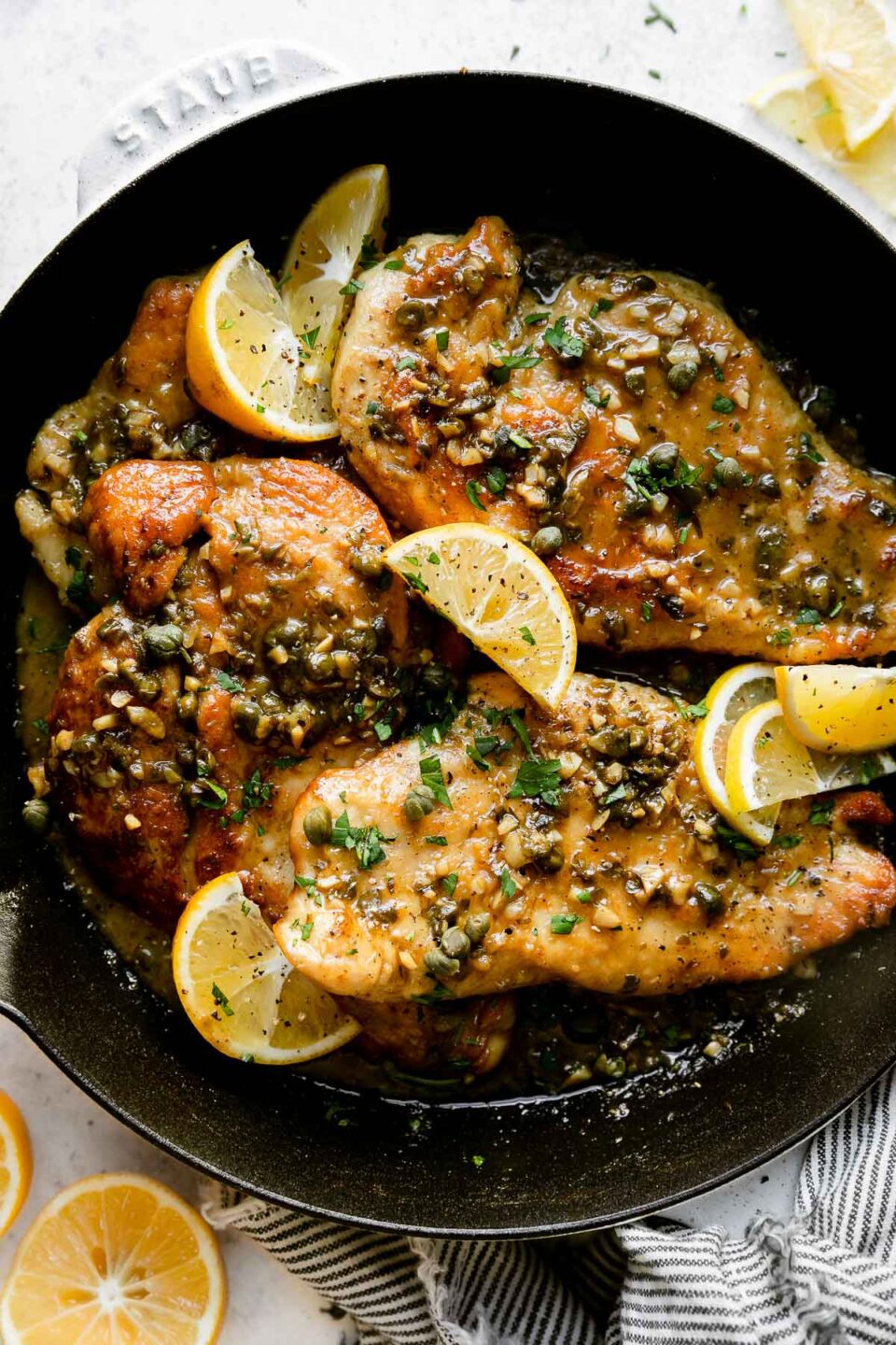 A white Staub cast iron skillet sits atop a creamy white textured surface filled with finished Meyer Lemon Chicken Piccata. The chicken cutlets have had Meyer Lemon pan sauce spooned on top and garnished with chopped fresh herbs and Meyer Lemon wedges. Halved and quartered lemons, loose fresh herbs, and a white and blue linen napkin surround the skillet.