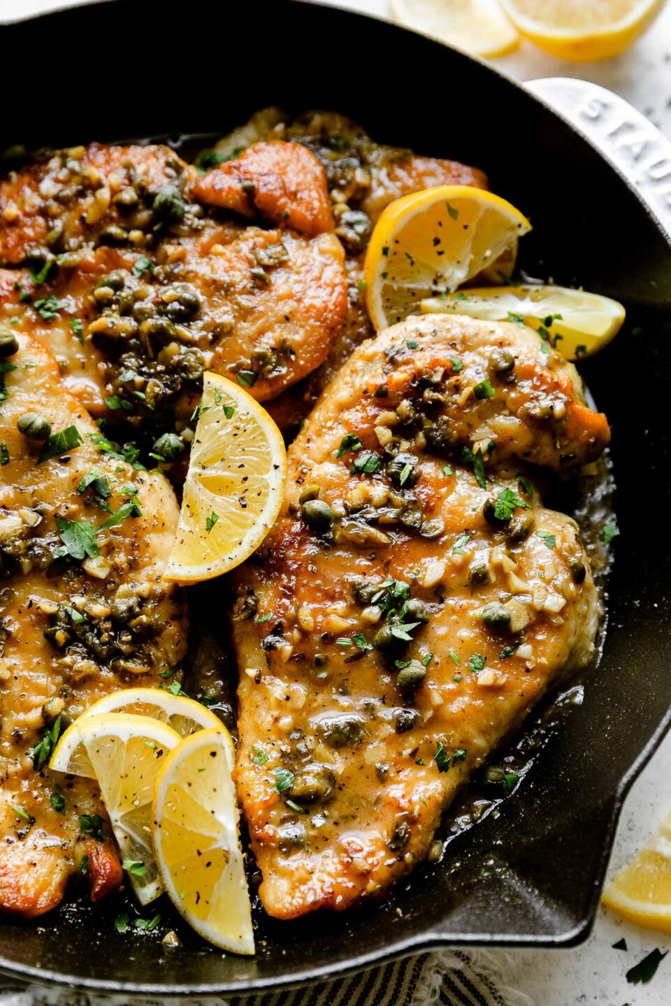 A close up shot of a white Staub cast iron skillet sits atop a creamy white textured surface filled with finished Meyer Lemon Chicken Piccata. The chicken cutlets have Meyer Lemon pan sauce spooned on top and garnished with chopped fresh herbs and Meyer Lemon wedges. Lemon wedges and a white and blue linen napkin surround the skillet.