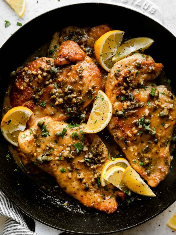 An overhead shot of a white Staub cast iron skillet sits atop a creamy white textured surface filled with finished Meyer Lemon Chicken Piccata. The chicken cutlets have Meyer Lemon pan sauce spooned on top and garnished with chopped fresh herbs and Meyer Lemon wedges. Lemon wedges and a white and blue linen napkin surround the skillet.