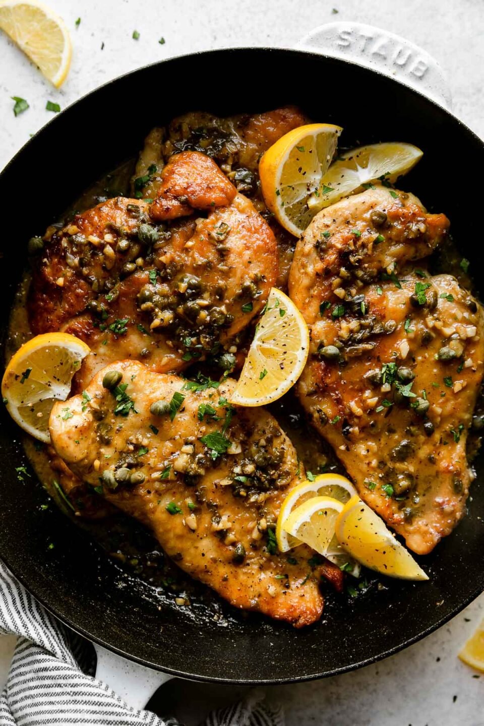 An overhead shot of a white Staub cast iron skillet sits atop a creamy white textured surface filled with finished Meyer Lemon Chicken Piccata. The chicken cutlets have Meyer Lemon pan sauce spooned on top and garnished with chopped fresh herbs and Meyer Lemon wedges. Lemon wedges and a white and blue linen napkin surround the skillet.