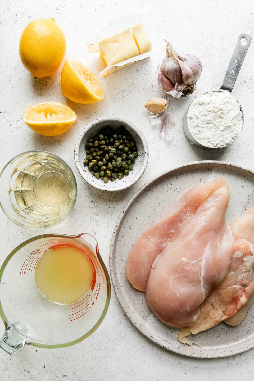 Meyer Lemon Chicken Piccata ingredients arranged on a creamy white textured surface: boneless, skinless chicken breasts, all-purpose flour, olive oil, garlic, capers, Meyer lemons, dry white wine, and unsalted butter.