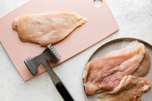 Butterflied chicken thats been pounded with a meat tenderizer rests atop a grey ceramic plate. A single butterflied chicken cutlet lays flat on a light pink cutting board next to the plate with a metal meat tenderizer resting atop the cutting board.